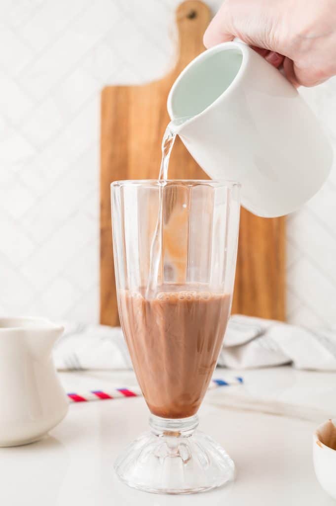 A hand pours milk from a white pitcher into a glass of iced coffee on a kitchen counter, reminiscent of crafting an Egg Cream Soda with its creamy swirl.