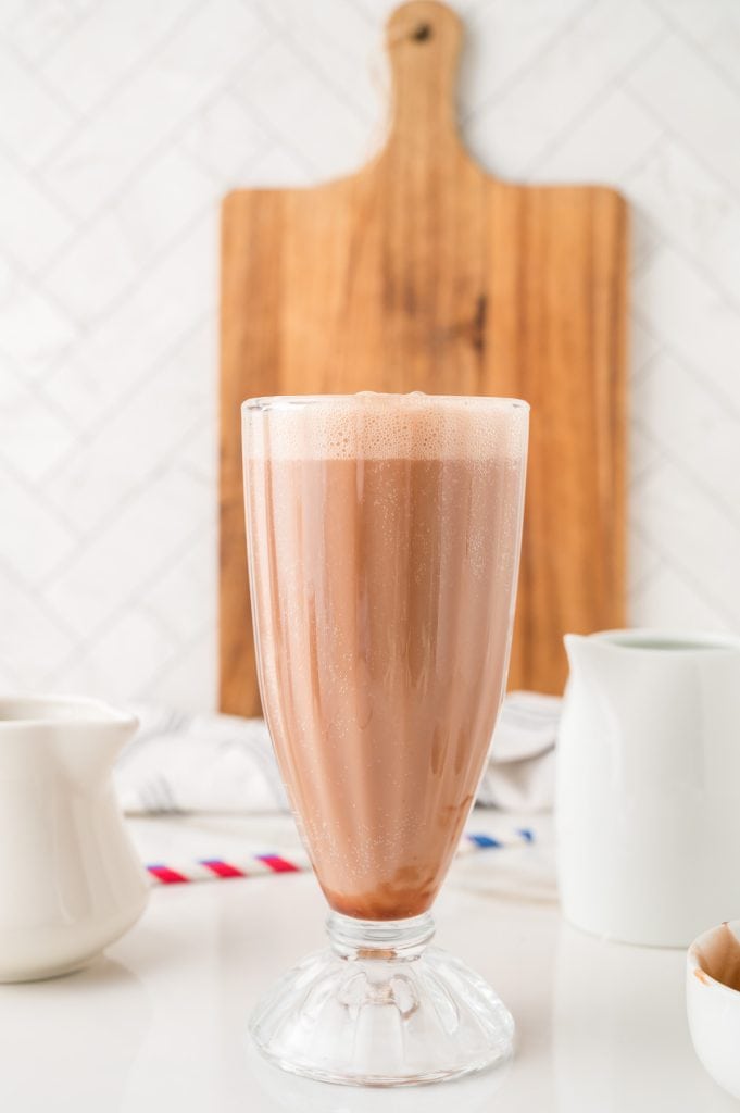 A tall glass of chocolate milkshake on a white counter with a wooden board in the background, reminiscent of a classic Egg Cream Soda enjoyed at an old-fashioned soda fountain.