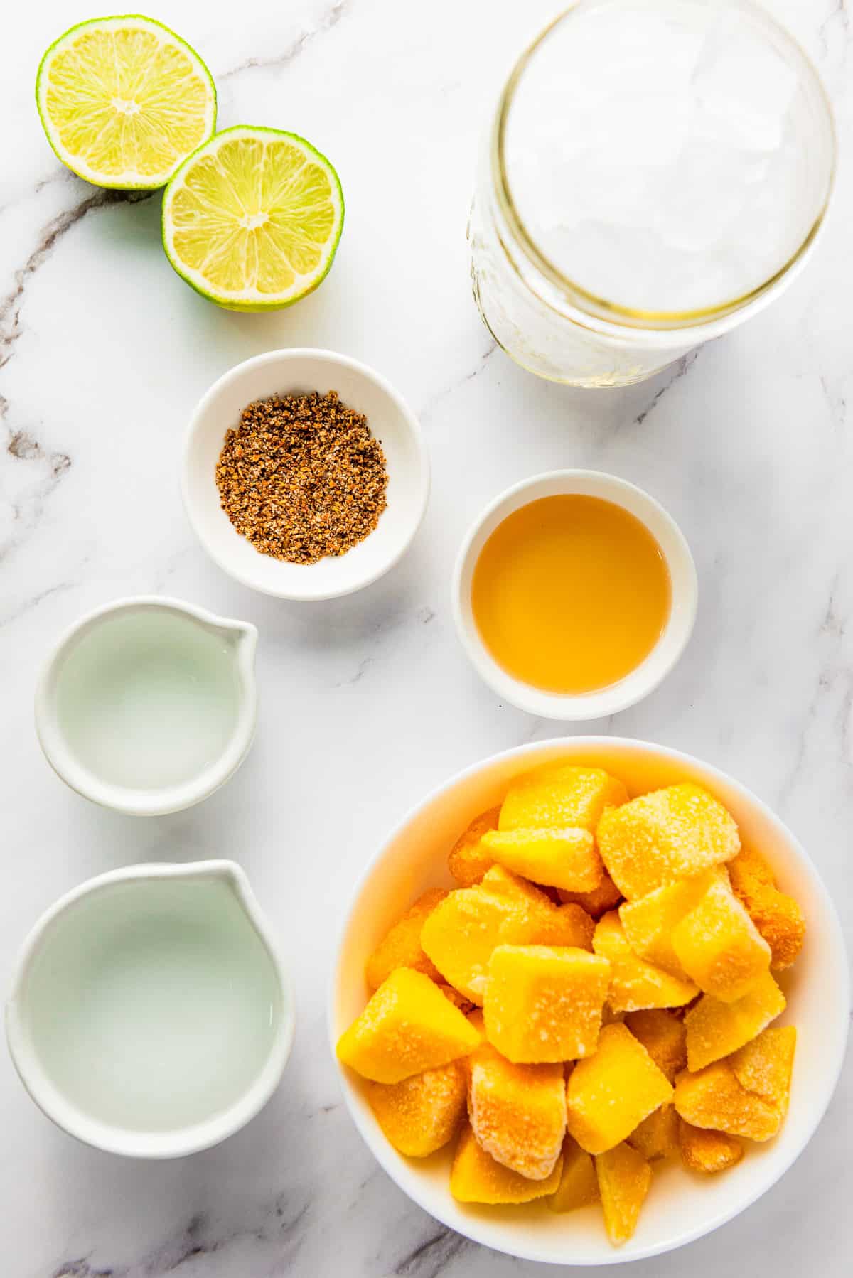 Frozen mango chunks, sliced lime, spices, honey, two liquids, and a glass of ice on a marble surfaceโperfect ingredients for crafting a refreshing frozen mango margarita mocktail.