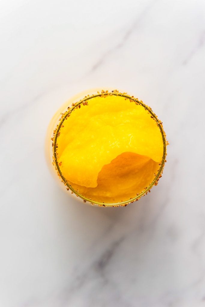 Top view of a yellow frozen mango margarita mocktail in a glass with a spiced rim on a white marble surface.