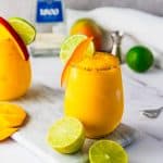Yellow frozen mango margarita mocktail with lime and chili rim, garnished with a lime slice, fresh mango, and a bottle in the background.
