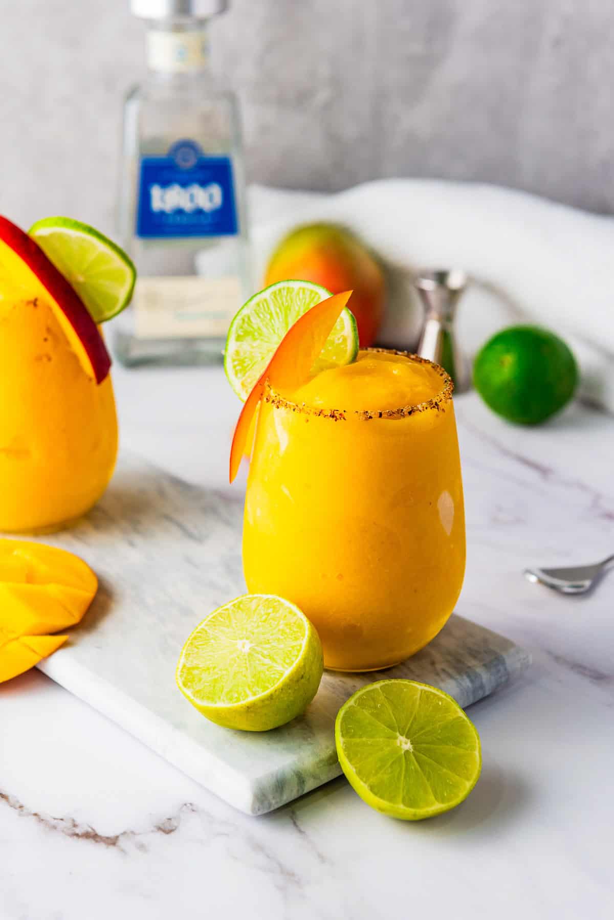 Yellow frozen mango margarita mocktail with lime and chili rim, garnished with a lime slice, fresh mango, and a bottle in the background.