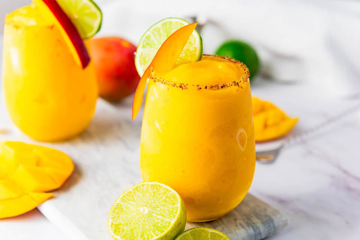 A frozen mango margarita mocktail in a glass with lime and mango slices, rimmed with chili powder, on a marble surface.
