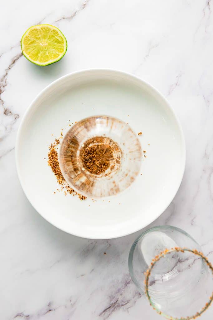 A glass with a sugared rim lies beside a bowl of brown sugar and a halved lime on a marble surface, ready to be transformed into a refreshing frozen mango margarita mocktail.