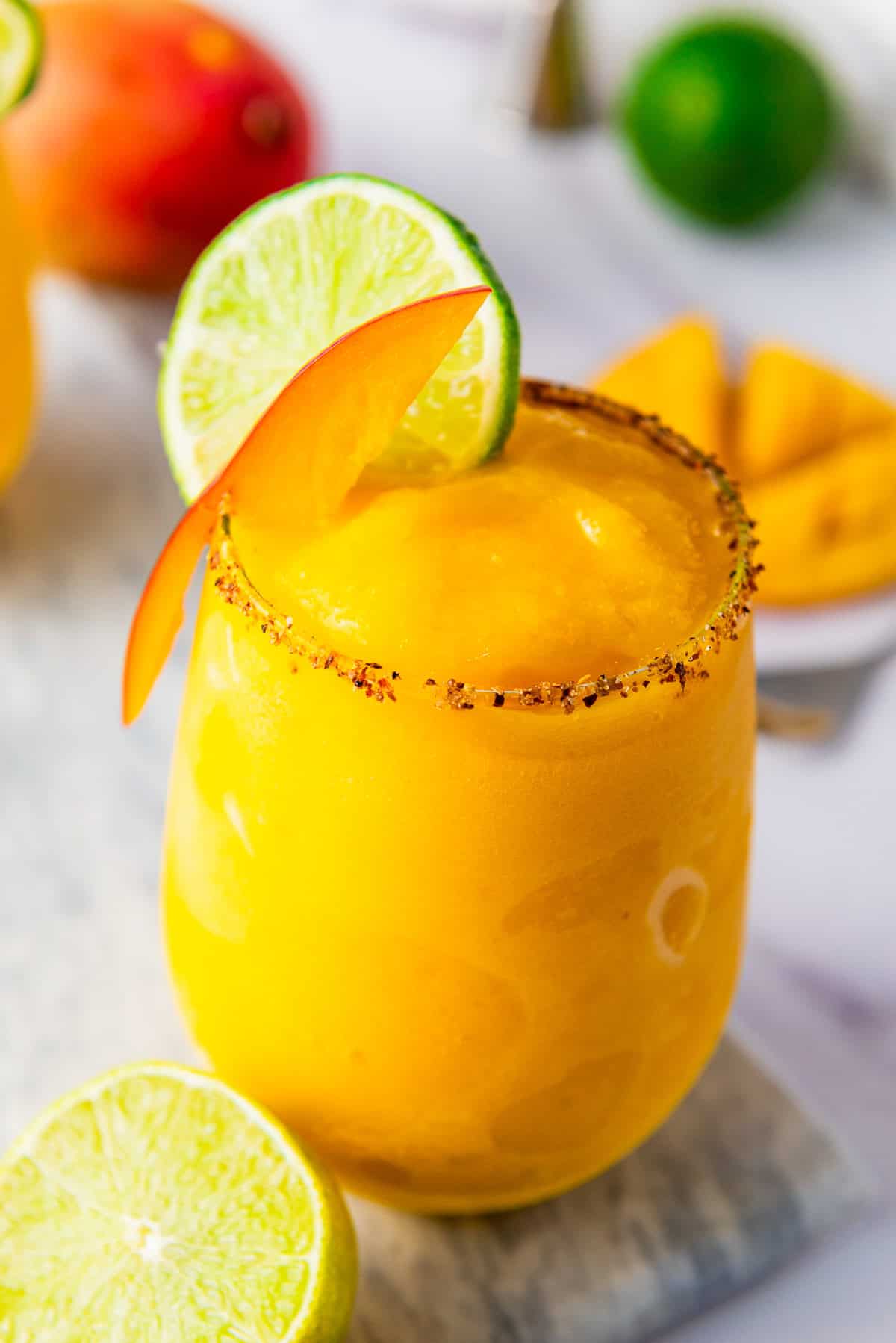 Bright yellow frozen mango margarita mocktail in a glass, garnished with lime, mango slice, and a chili rim.