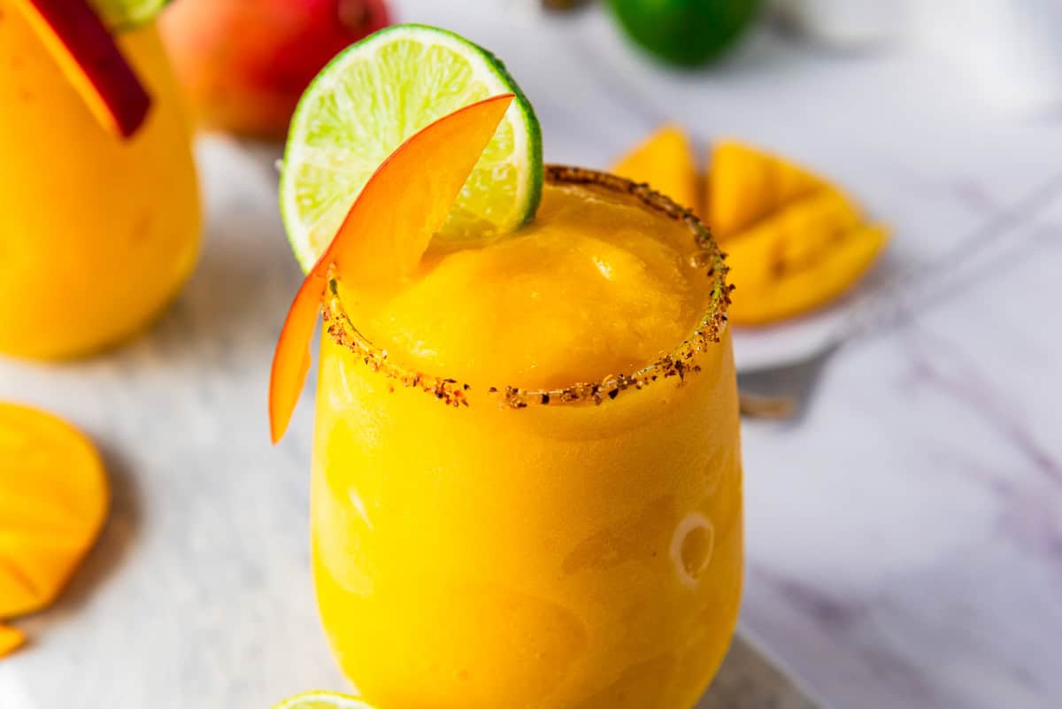 A yellow frozen mango margarita mocktail in a glass, garnished with lime, mango slice, and chili powder on the rim.