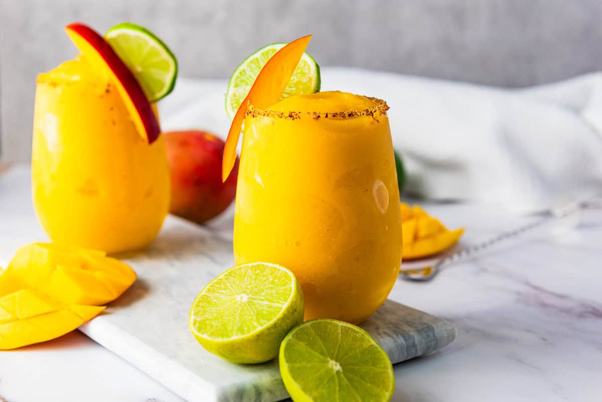 Two frozen mango margarita mocktails with lime and mango slices, garnished with chili on the rim, on a marble board.
