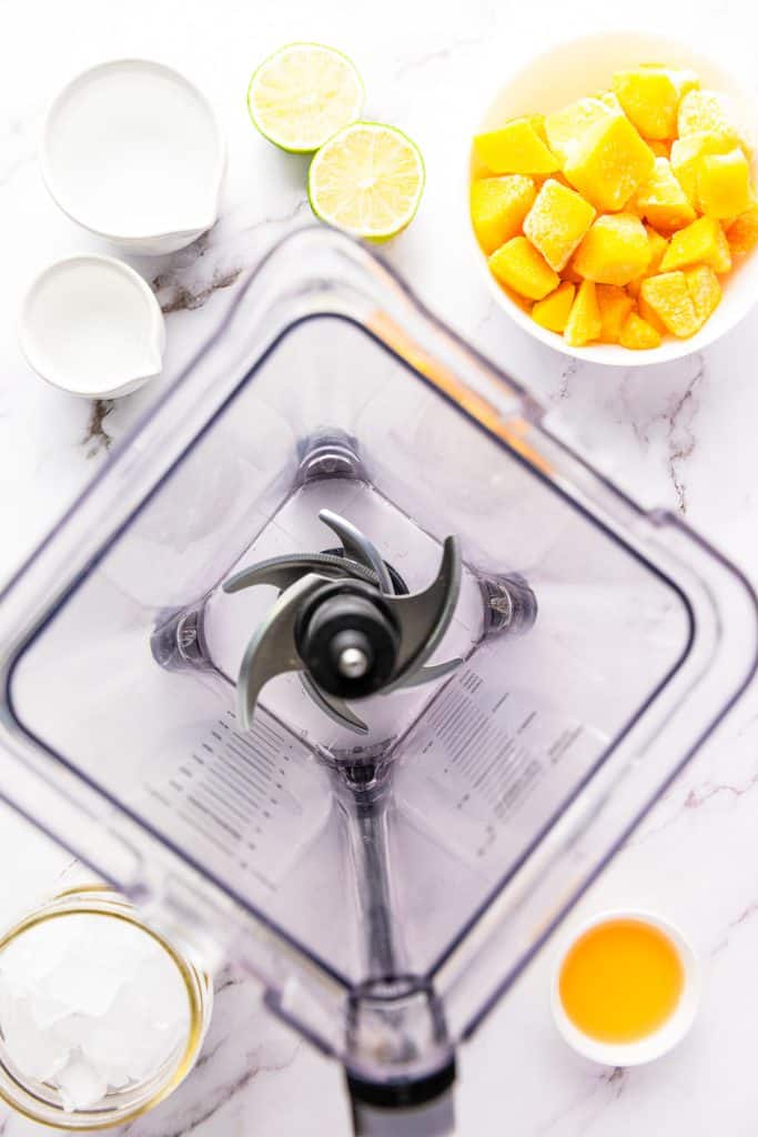 Top view of a blender surrounded by ingredients for a frozen mango margarita mocktail, including fresh mango, lime, orange juice, coconut milk, and ice on a marble surface.