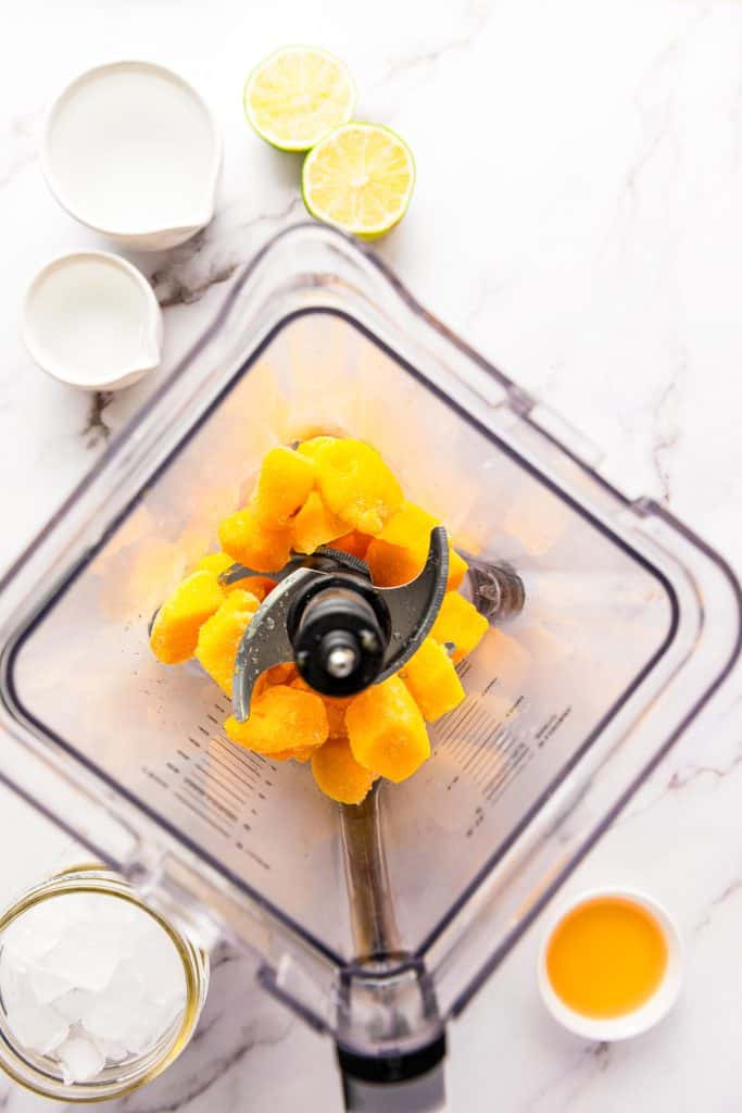 Top view of a blender with frozen mango chunks, surrounded by lime, milk, juice, and a cup of ice on a counterโperfect ingredients for crafting a refreshing frozen mango margarita mocktail.