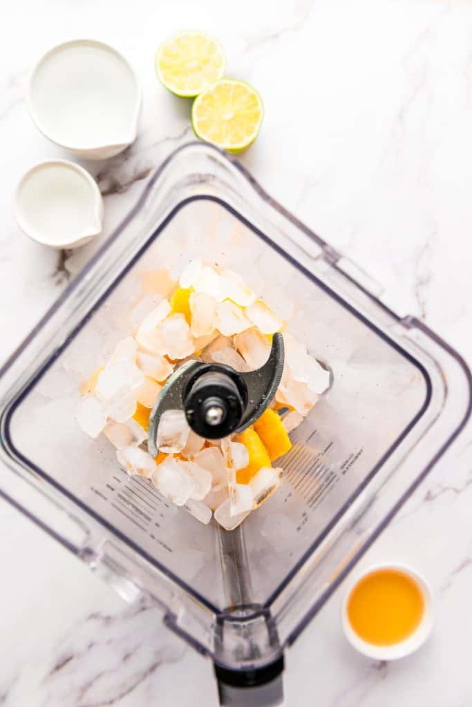 Top view of a blender with ice and lemon, perfect for preparing a frozen mango margarita mocktail, surrounded by lime halves, milk, cream, and orange juice on a white table.
