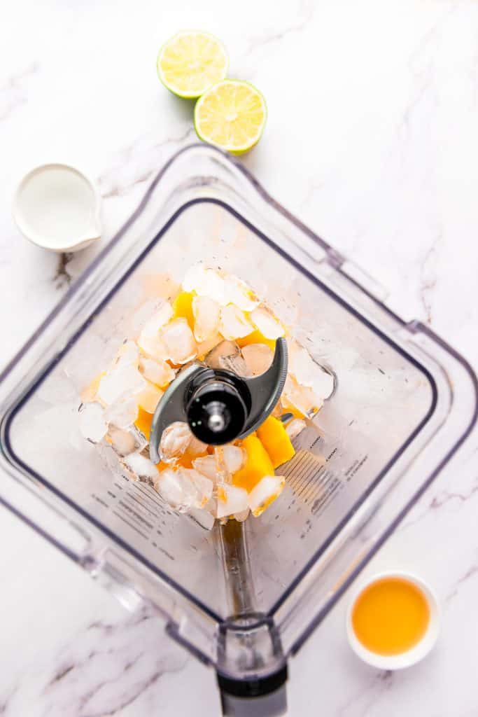 Top view of a blender with ice and mango, ready to be blended into a delicious frozen mango margarita mocktail, surrounded by lime, juice, and milk on a marble counter.
