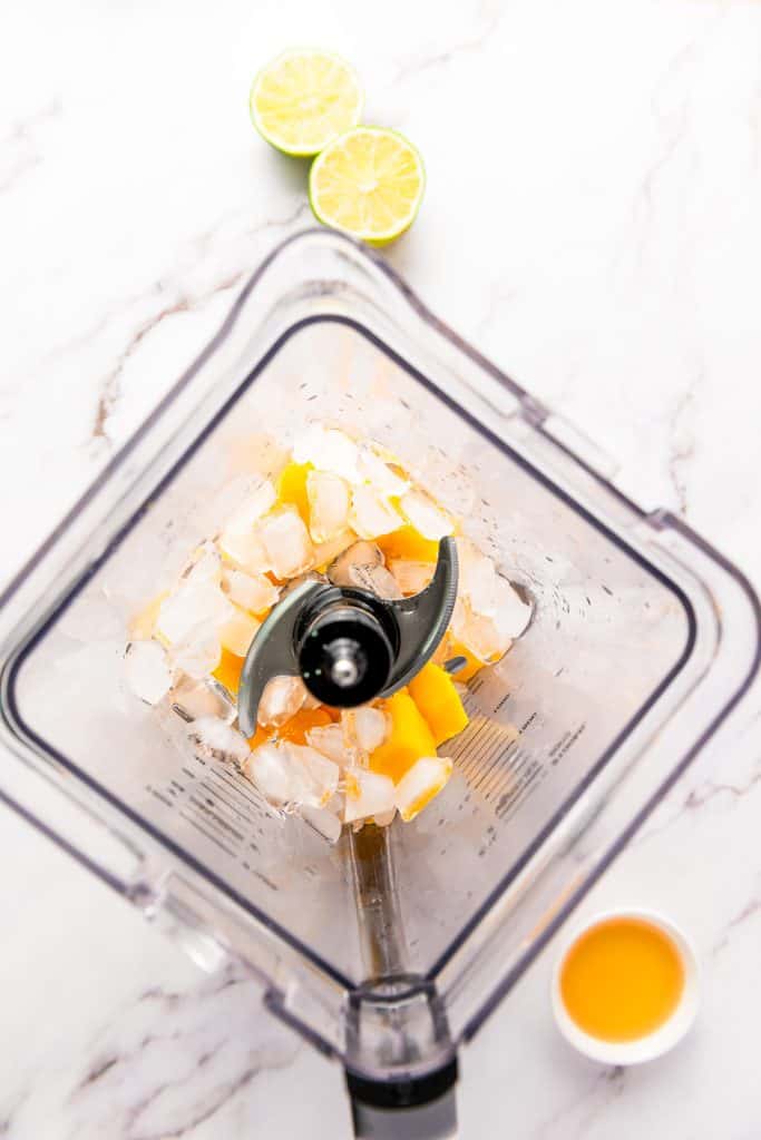 Chunks of mango and ice in a blender, with halved limes and a small cup of juice nearby on a white surfaceโperfect ingredients for crafting a refreshing frozen mango margarita mocktail.