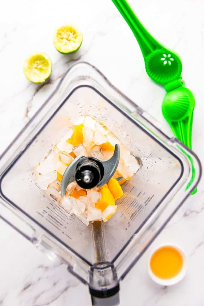 Ice cubes and mango chunks in a blender, ready to become a refreshing frozen mango margarita mocktail, with a lime squeezer, halved limes, and fresh juice on a white surface.