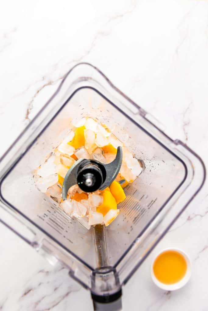 Top view of a blender with ice and mango chunks inside, perfect for creating a delicious frozen mango margarita mocktail, next to a small cup of orange liquid on a white surface.