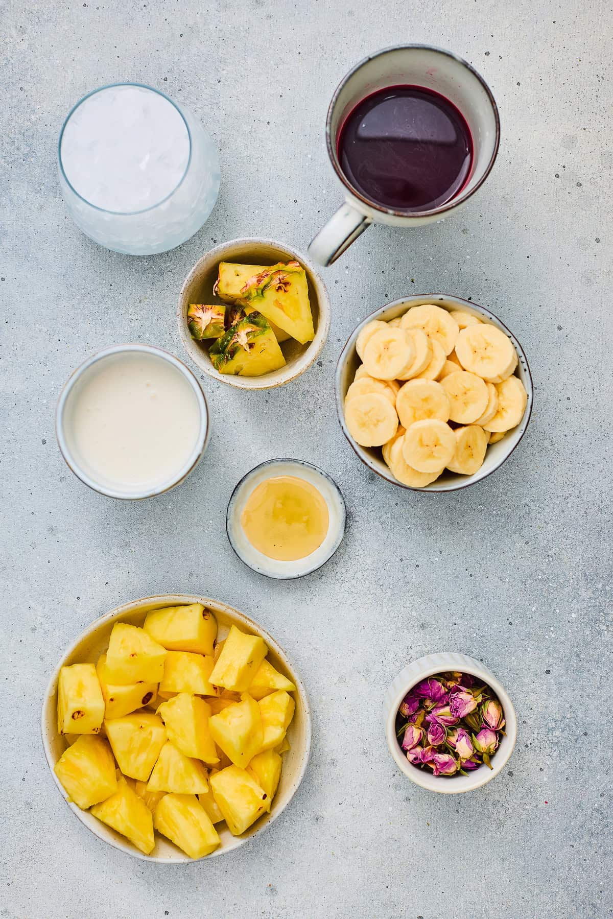 Bowls of pineapple, banana, honey, yogurt, rosebuds, a cup of juice, and a glass of milk on a gray surface create the perfect setup for making a refreshing Hibiscus Tea Pineapple Smoothie.