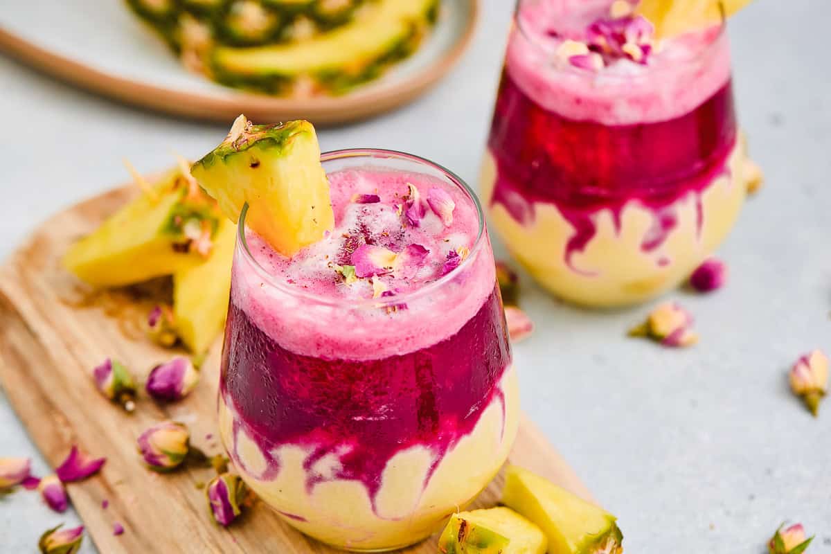 Two colorful layered Hibiscus Tea Pineapple Smoothies with pineapple wedges and rose petals, surrounded by pineapple chunks.