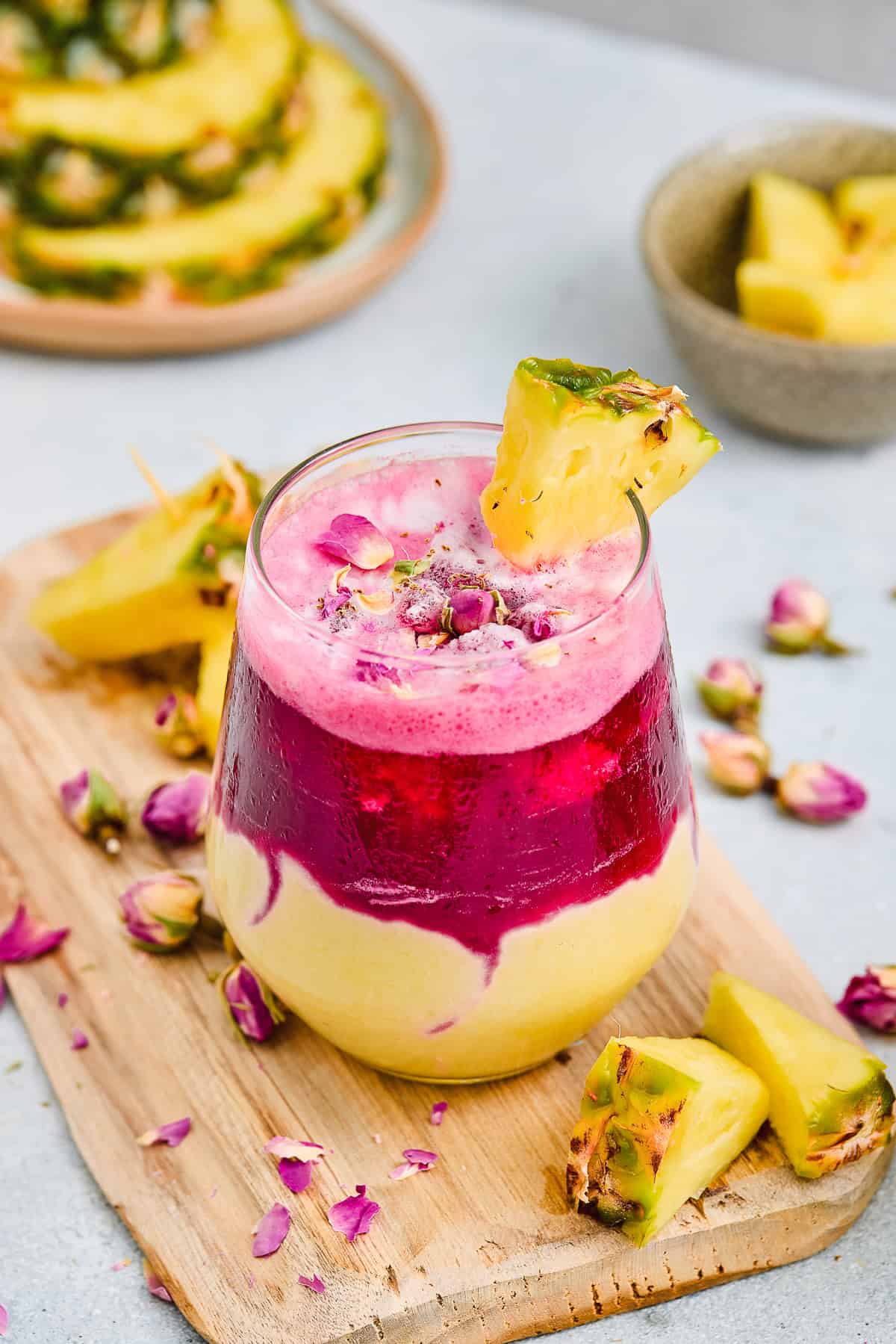 A layered Hibiscus Tea Pineapple Smoothie with yellow and pink swirls, garnished with pineapple and rose petals on a wooden board.