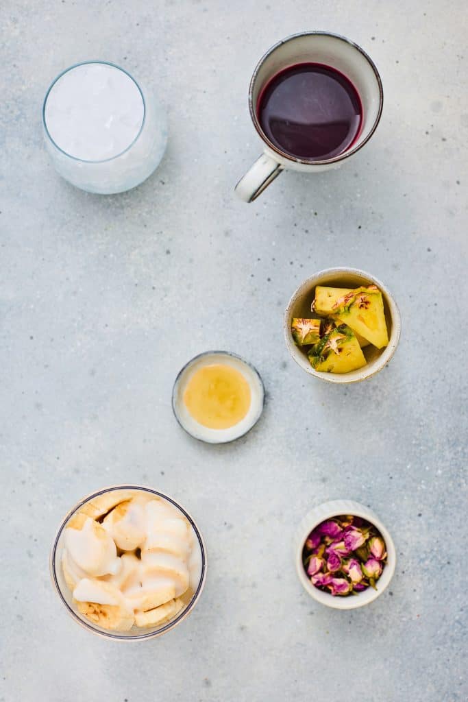 Flat lay of assorted drinks and snacks in cups and bowls on a light surface, including fruit, ice, and petals, featuring vibrant Hibiscus Tea and refreshing Pineapple Smoothie.