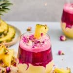 Layered Pineapple Smoothie with bright pink and yellow swirls, topped with a pineapple slice and dried rose petals. The vibrant colors are enhanced by a refreshing Hibiscus Tea layer, making this smoothie as beautiful as it is delicious.