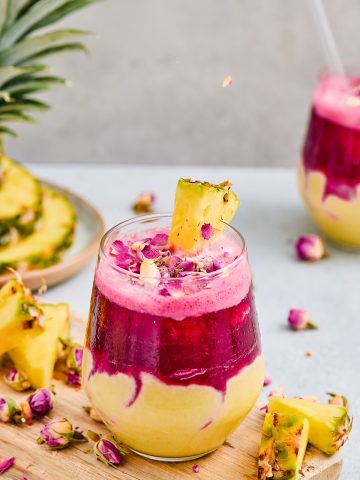 Layered Pineapple Smoothie with bright pink and yellow swirls, topped with a pineapple slice and dried rose petals. The vibrant colors are enhanced by a refreshing Hibiscus Tea layer, making this smoothie as beautiful as it is delicious.