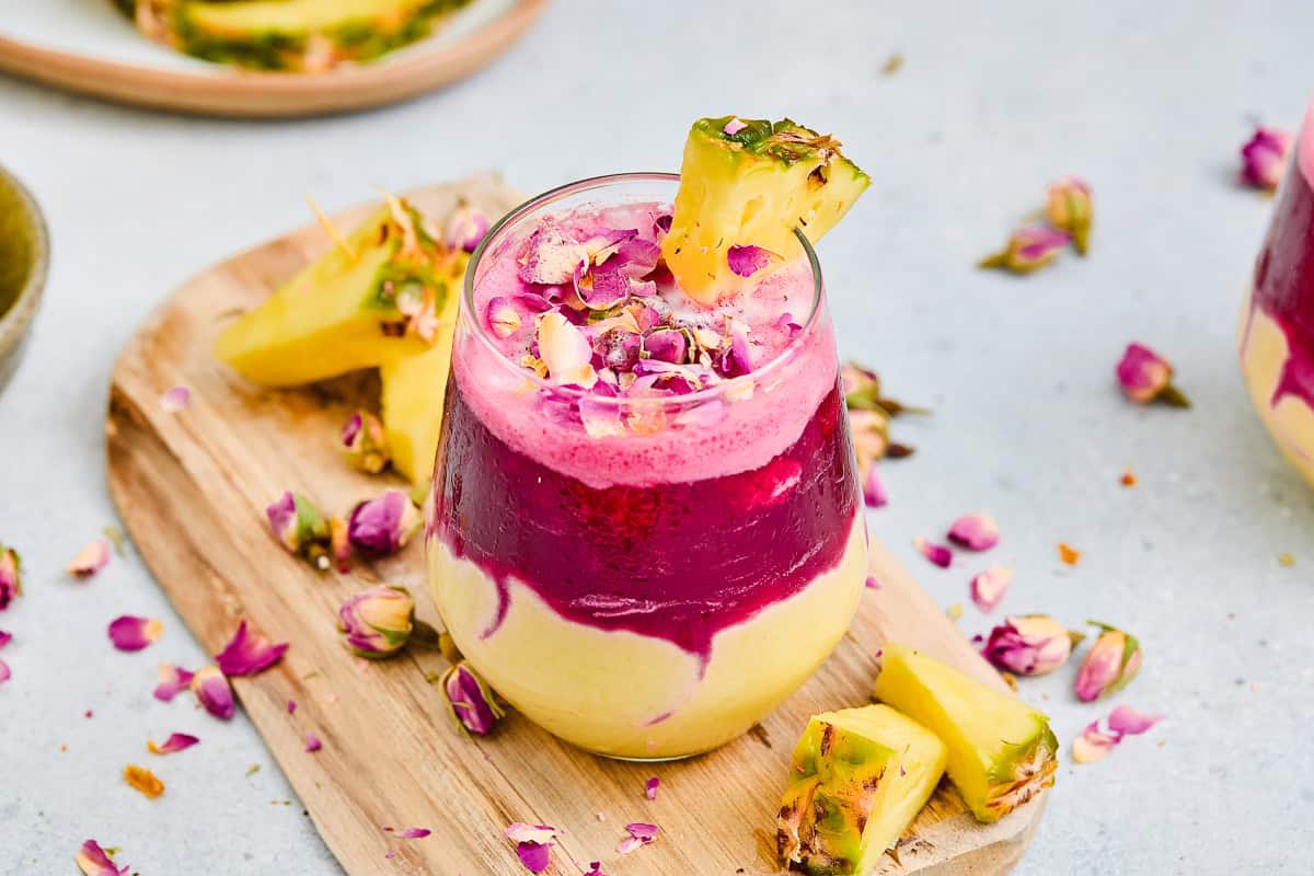 A layered Hibiscus Tea Pineapple Smoothie with vibrant pink and yellow colors, garnished with pineapple and rose petals on a wooden board.