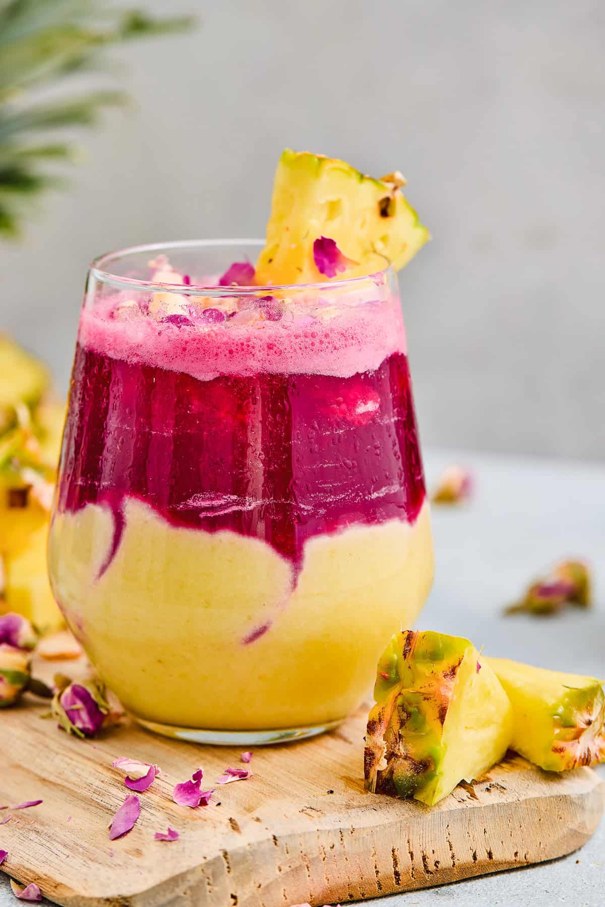 A vibrant Hibiscus Tea Pineapple Smoothie featuring yellow and pink layers, elegantly garnished with a pineapple wedge, served on a wooden board.