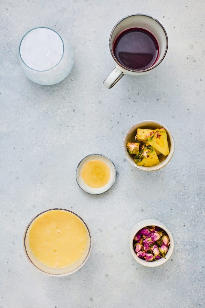 Six cups with ice, red hibiscus tea, yellow pineapple smoothie, pineapple pieces, rose buds, and a small cup of light liquid sit on a gray surface.