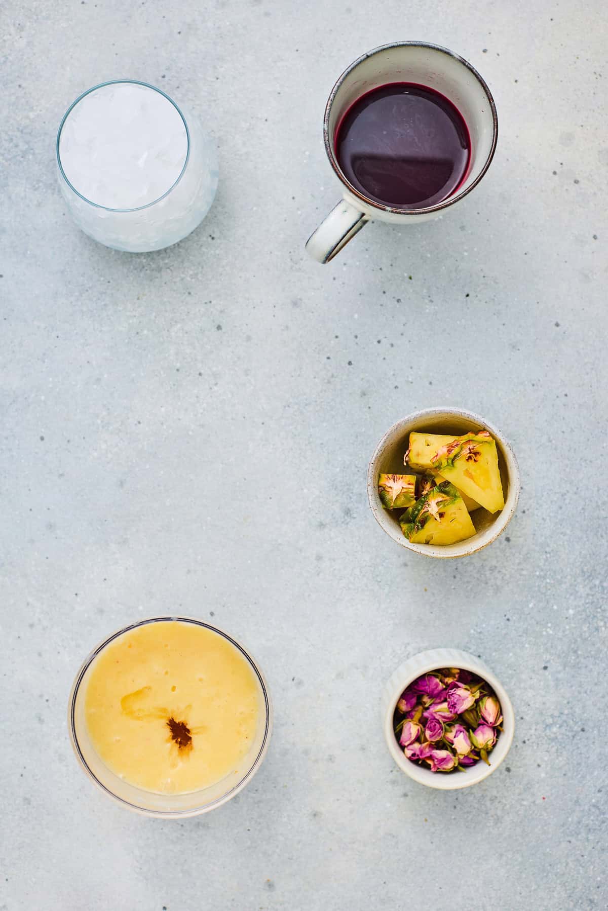 Five cups on a light surface, filled with ice, refreshing hibiscus tea, red liquid, pineapple smoothie, yellow drink, and dried rosebuds.