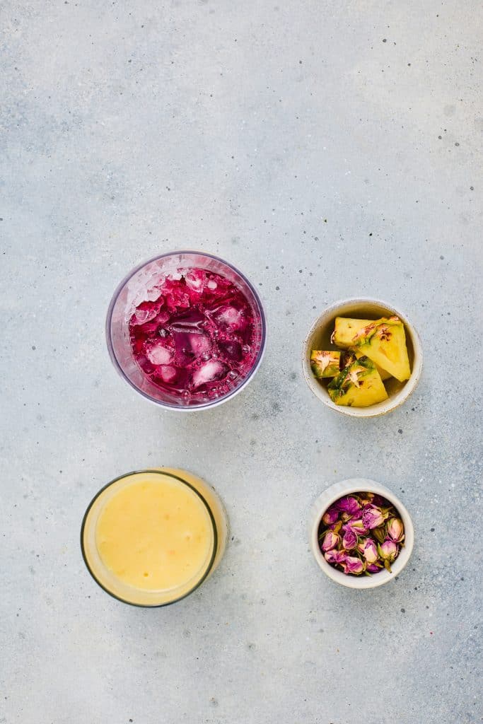 A flat lay of a vibrant Hibiscus Tea, a refreshing Pineapple Smoothie, pineapple chunks, and dried pink rosebuds on a gray surface.