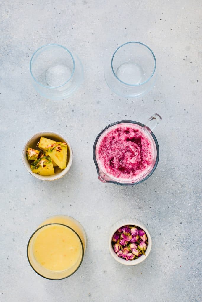 Two empty glasses, a jug of Hibiscus Tea Pineapple Smoothie, yellow smoothie, pineapple slices, and dried rosebuds on a light surface.