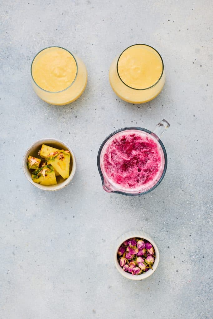 Two glasses of yellow Hibiscus Tea Pineapple Smoothie, pineapple chunks, a pink smoothie in a jug, and a bowl of dried rosebuds.