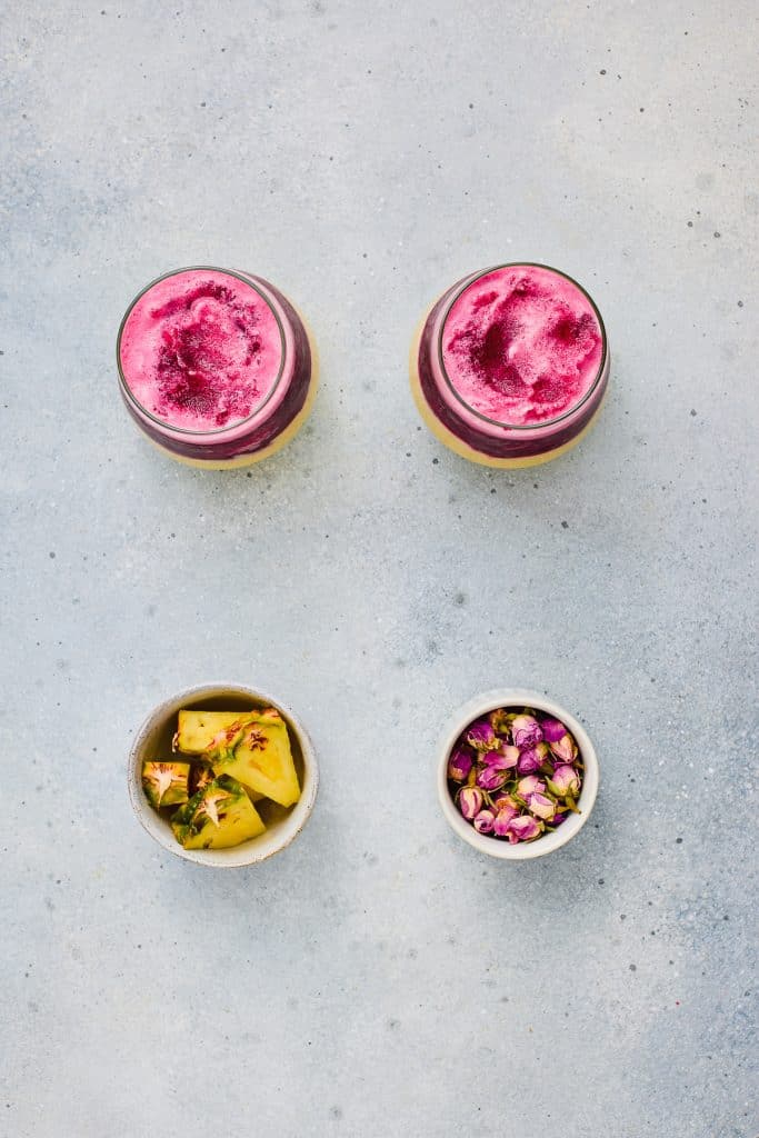 Two pink Hibiscus Tea Pineapple Smoothies in glasses, with bowls of pineapple chunks and dried rosebuds on a light gray surface.
