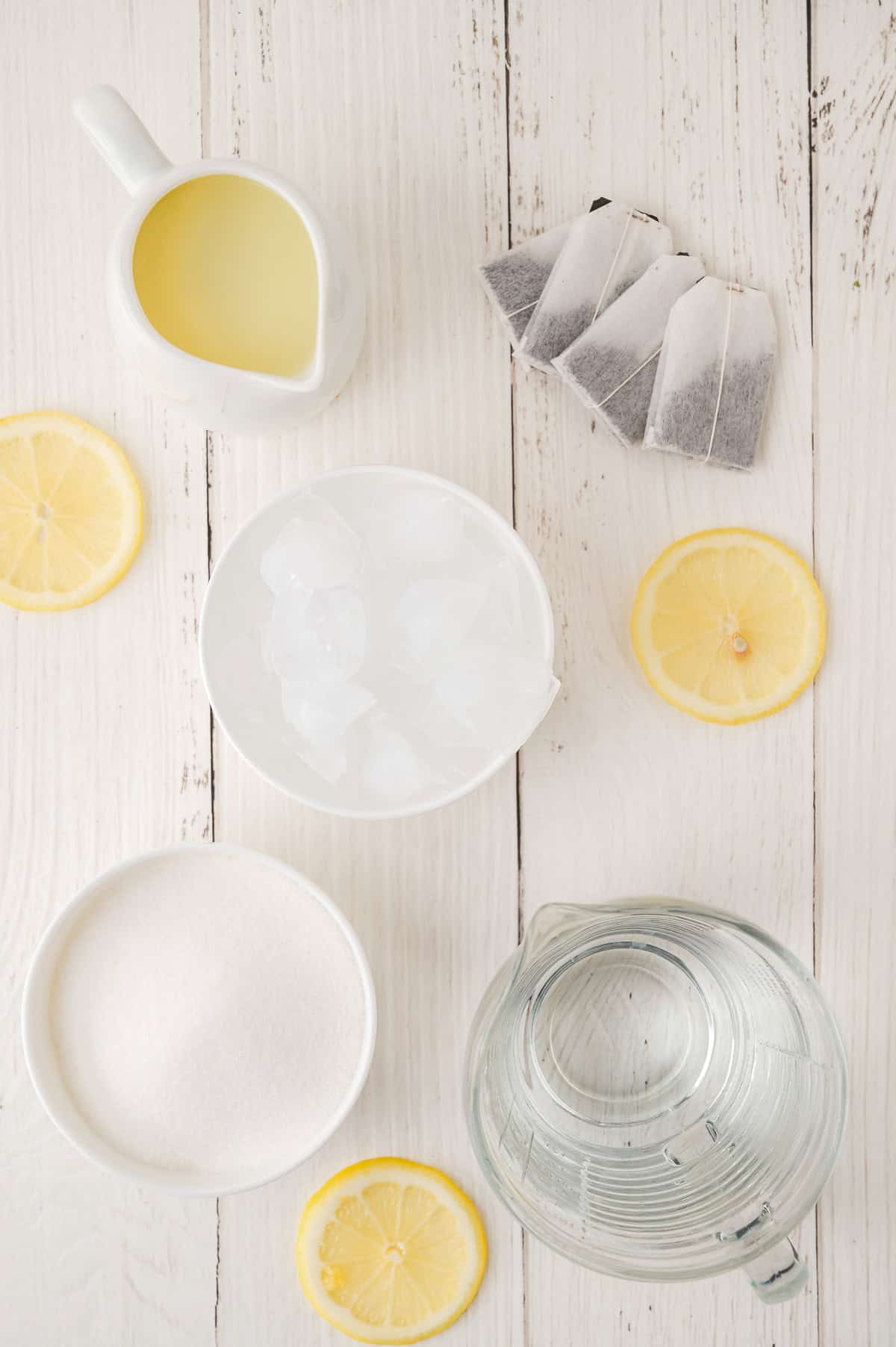 Top view of tea bags, ice, sugar, lemon slices, water, and a pitcher with a yellow liquid—perfect ingredients for making refreshing Lemonade Iced Tea—arranged on white wood.
