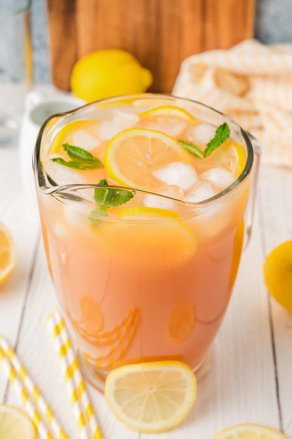 A glass pitcher of refreshing Lemonade Iced Tea with lemon slices and mint, on a white table with yellow striped straws.