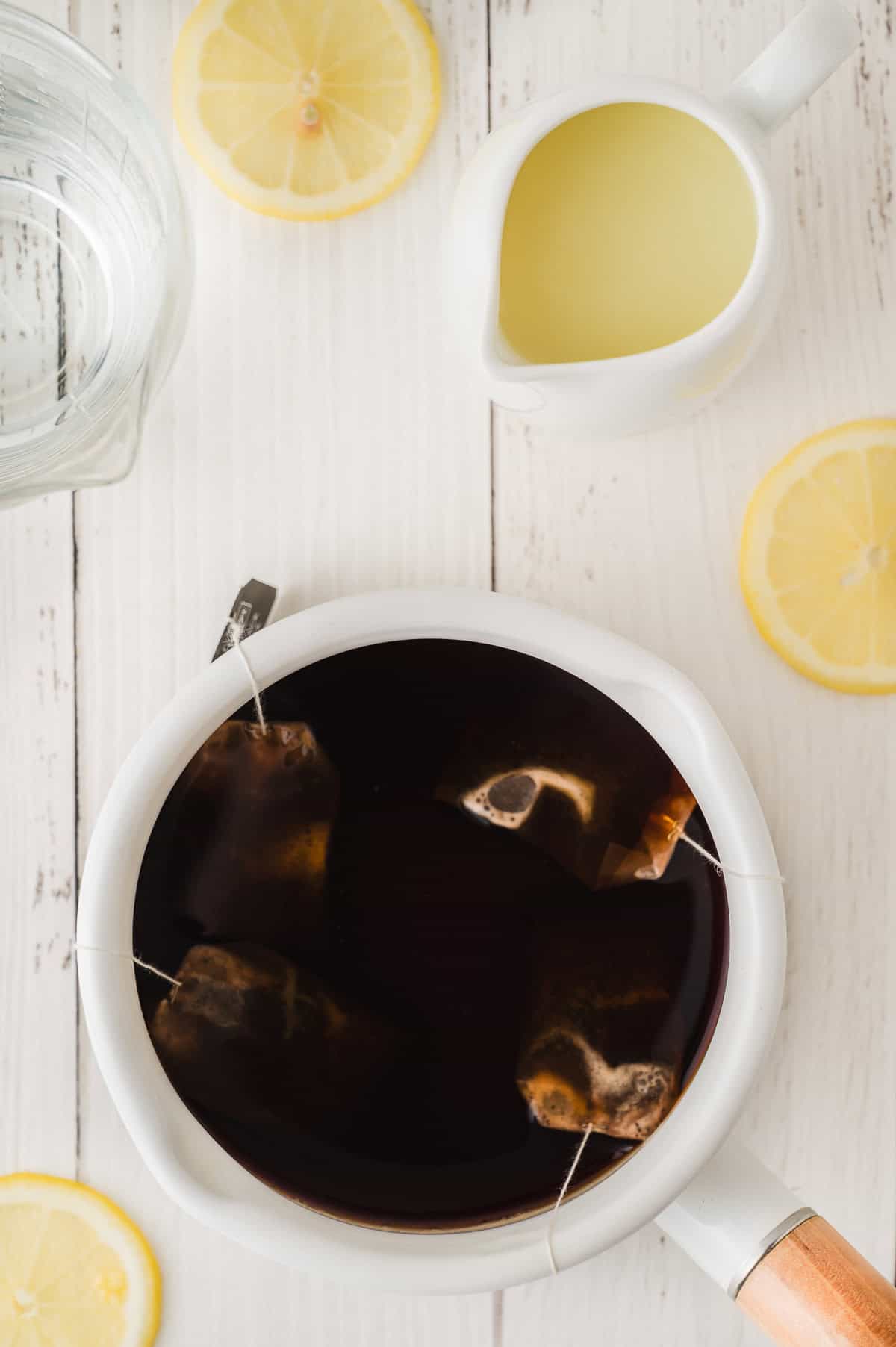 A white pot with tea bags in hot water, surrounded by lemon slices and a small pitcher of lemon juice, perfectly captures the refreshing spirit of Lemonade Iced Tea.