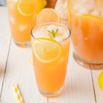 Lemonade Iced Tea with lemon slices and mint served in two glasses and a pitcher on a white table with a wooden board.