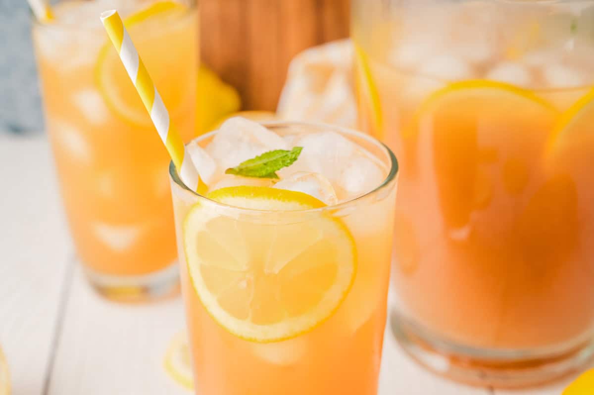 A glass of iced lemonade with a lemon slice, mint, and a striped straw, next to a pitcher&mdash;perfect for enjoying classic Lemonade Iced Tea on a warm day.