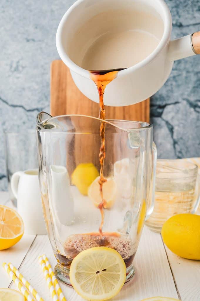 Coffee is being poured from a white pitcher into a glass pitcher with lemon slices on a table, creating a unique fusion reminiscent of Lemonade Iced Tea.