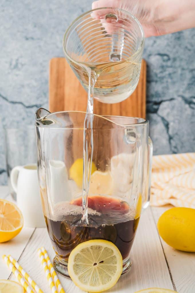 A hand pours clear liquid into a glass pitcher filled with dark Iced Tea, surrounded by a lemon slice and straws nearby.