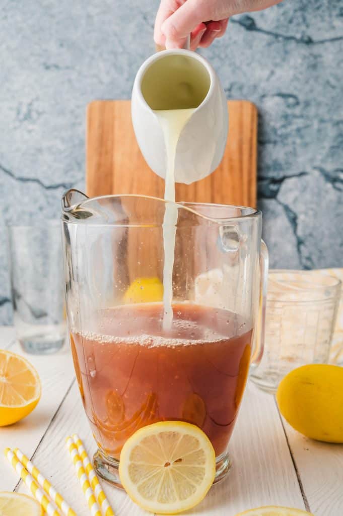 A hand pours lemon juice from a small white jug into a pitcher of Lemonade Iced Tea, with lemon slices nearby.