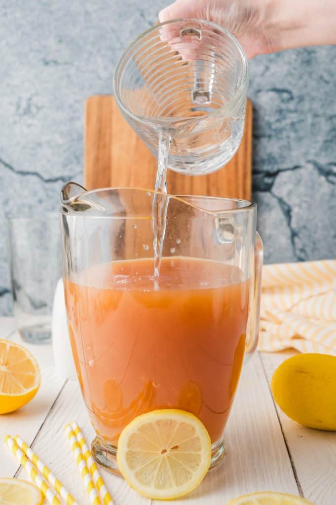 A hand pours water from a glass cup into a pitcher of lemonade iced tea, surrounded by lemons and straws nearby.