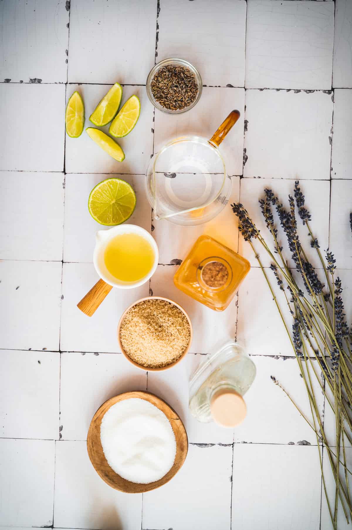 Lime wedges, lavender, seeds, sugar, honey, and liquids—perfect ingredients for crafting a refreshing lavender margarita mocktail—are artfully arranged on a white tiled surface.
