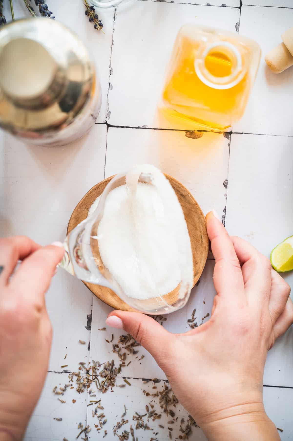 A hand dips a cocktail glass rim in sugar, surrounded by a shaker, liquor bottle, and scattered herbs, setting the stage for crafting a refreshing lavender margarita mocktail.