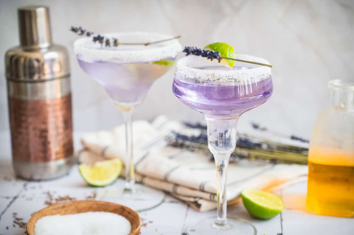 Two purple lavender margarita mocktail cocktails in sugar-rimmed glasses, garnished with lime and lavender, beside a cocktail shaker.