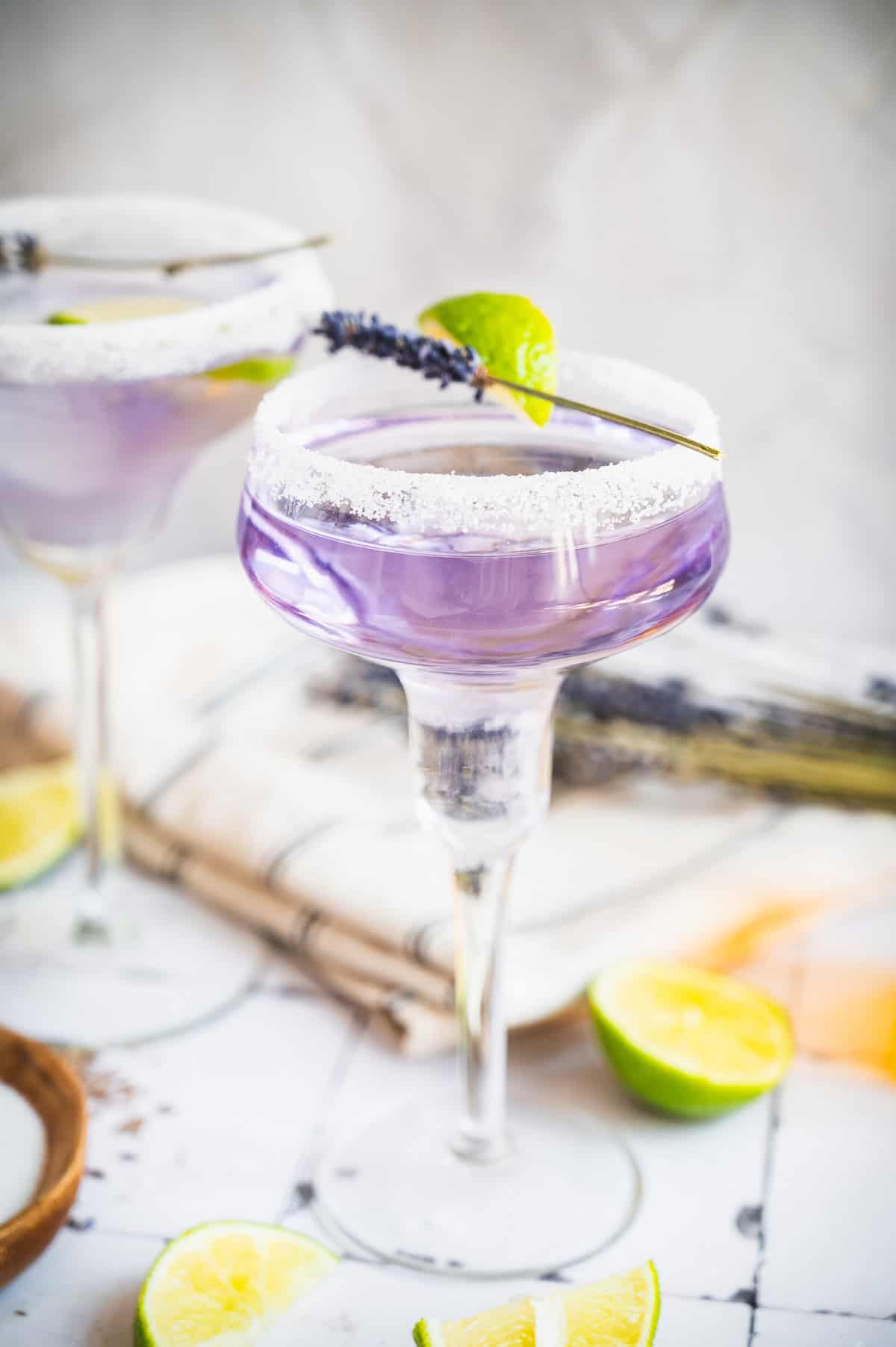 Purple lavender margarita mocktail served in a sugar-rimmed glass, garnished with lime and lavender, with lime slices nearby.