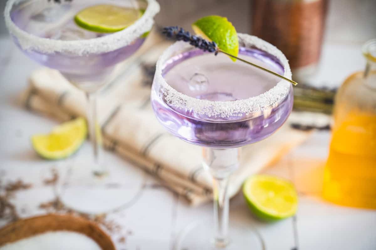 Purple lavender margarita mocktail in a sugar-rimmed glass, garnished with lime and lavender; lime wedges nearby.