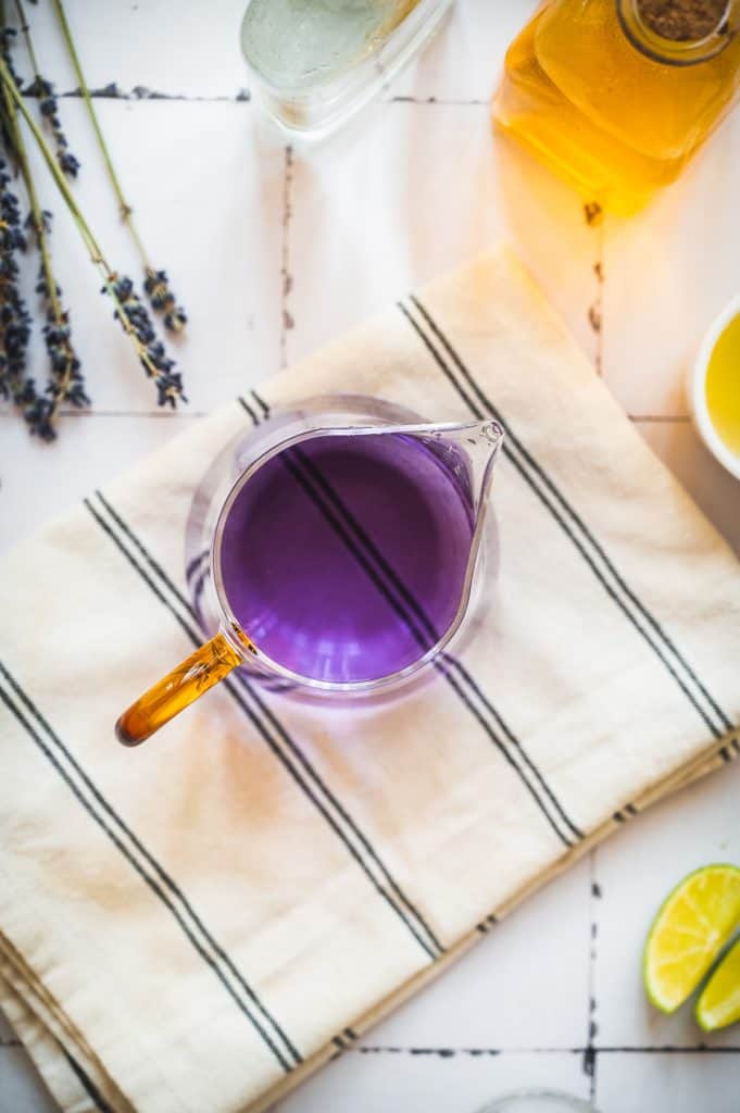 A glass pitcher with refreshing purple liquid, likely a lavender margarita mocktail, sits on a striped towel, surrounded by sprigs of lavender, zesty lime, and golden honey.