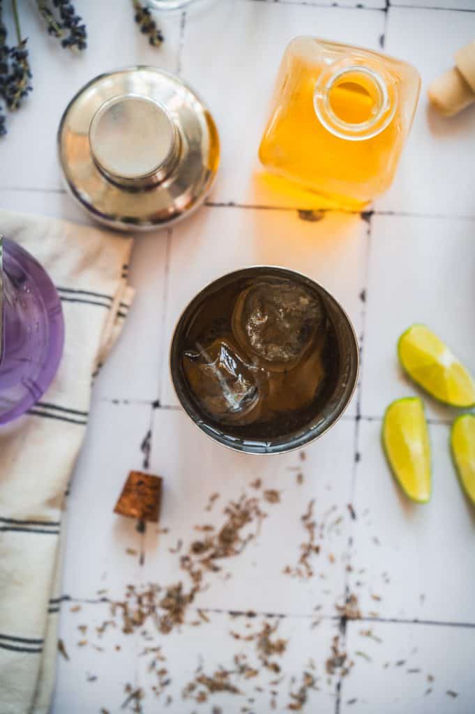 Overhead view of a lavender margarita mocktail with ice, lime wedges, shaker, and a jar with orange liquid on a tiled surface.