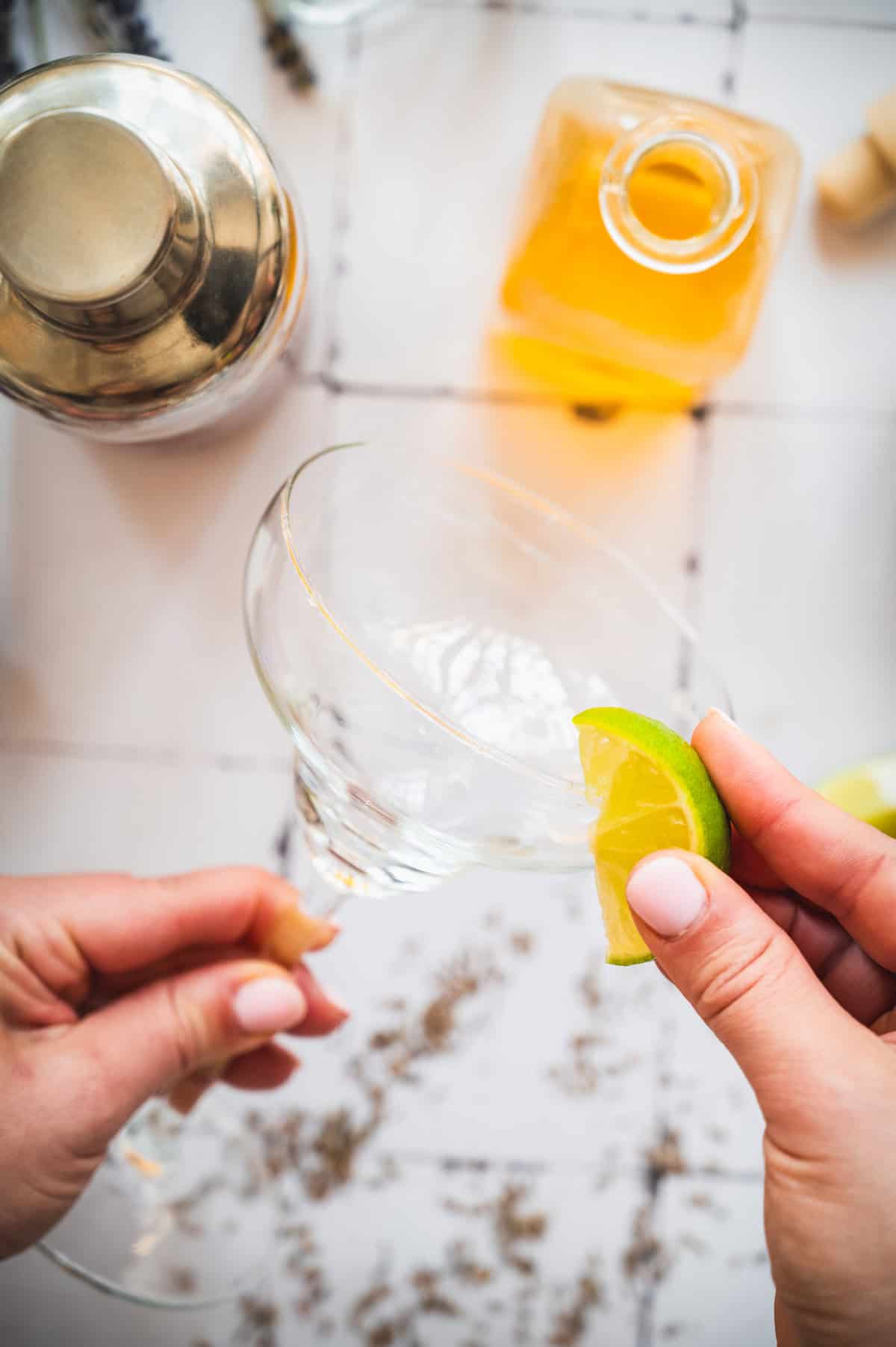 Hand rubbing a lime wedge on the rim of an empty cocktail glass, preparing the perfect base for a refreshing lavender margarita mocktail; shaker and liquor bottle nearby.