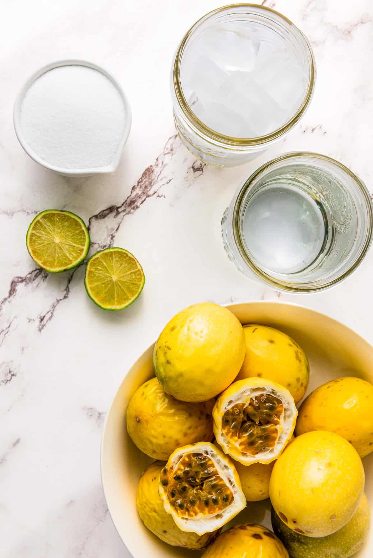 A bowl of passion fruits, cut limes, sugar, and ice water sit alongside two jars on a marble surface—perfect for making refreshing Passion Fruit Juice.